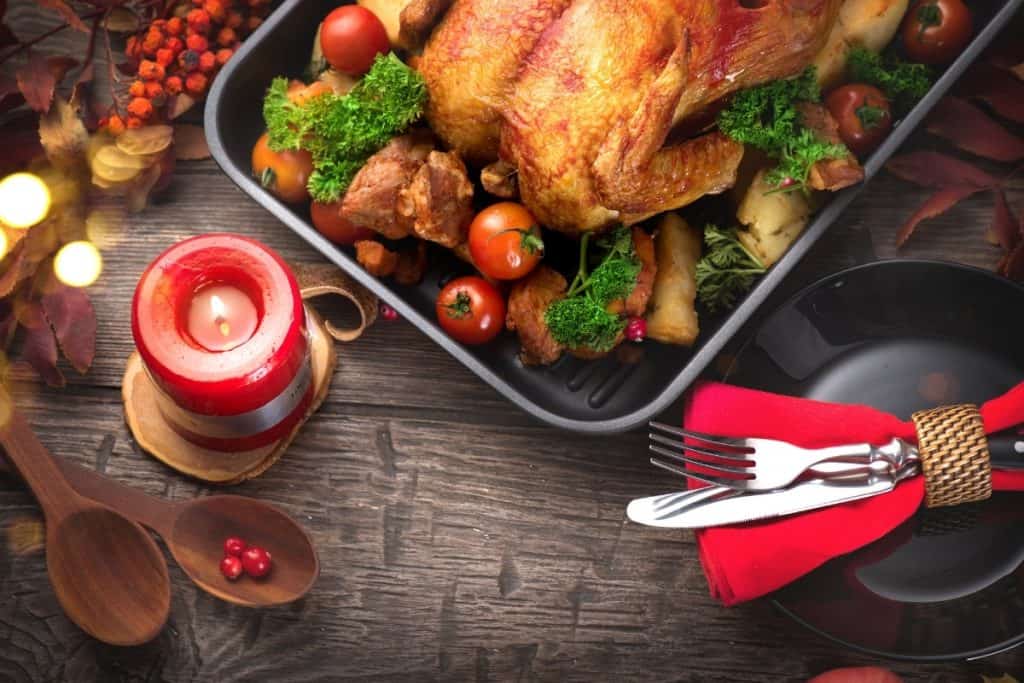 A close-up of a roasted chicken garnished with cherry tomatoes, parsley, and other vegetables in a baking dish. The setting includes a red candle and festive decorations on a rustic wooden table, creating a holiday ambiance.