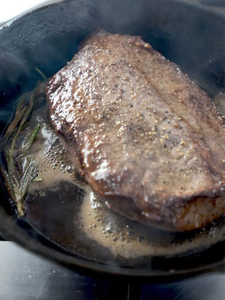 Flat iron steak searing in a cast iron skillet, with steam rising during the cooking process.