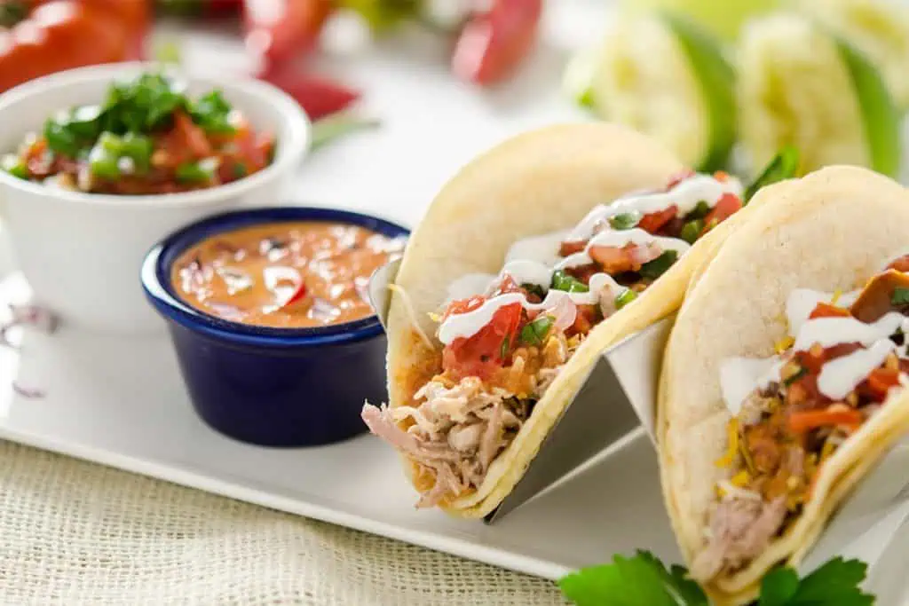 A close-up of two tacos on a plate. The tacos are filled with pulled pork barbacoa, topped with shredded lettuce, pico de gallo salsa, and sour cream.