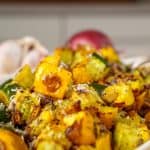 Roasted summer squash and zucchini served in a white dish, garnished with grated Parmesan cheese, with whole zucchini, yellow squash, red onion, and garlic surrounding the dish.