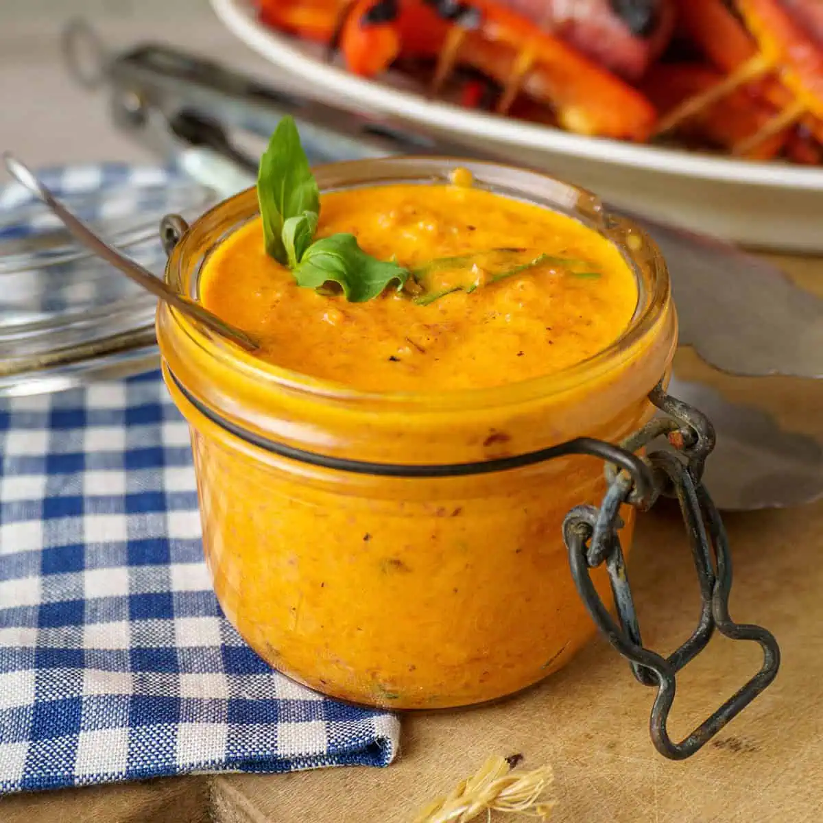 A close-up view of a glass jar filled with vibrant orange roasted red pepper sauce, garnished with fresh basil leaves. The jar is placed on a wooden board with a blue and white checkered napkin, and in the background, there is a plate of grilled vegetables.
