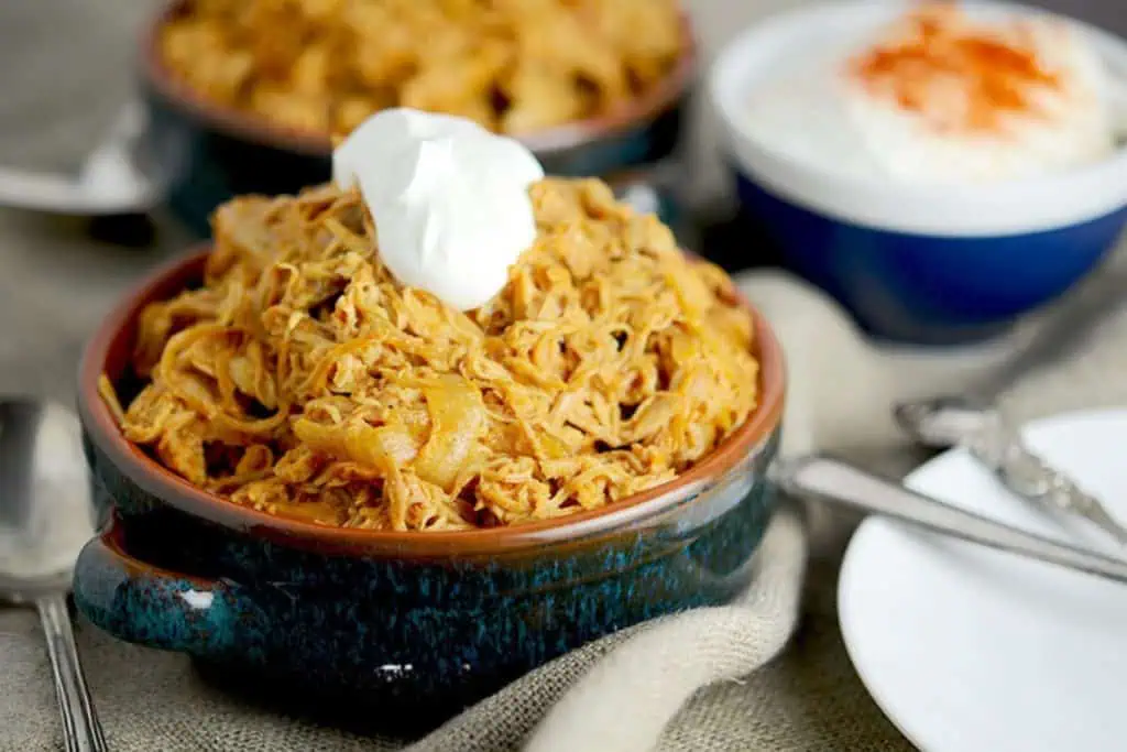 A bowl of shredded chicken paprikash garnished with sour cream, with a small bowl of paprika-spiced sour cream and a spoon in the background.