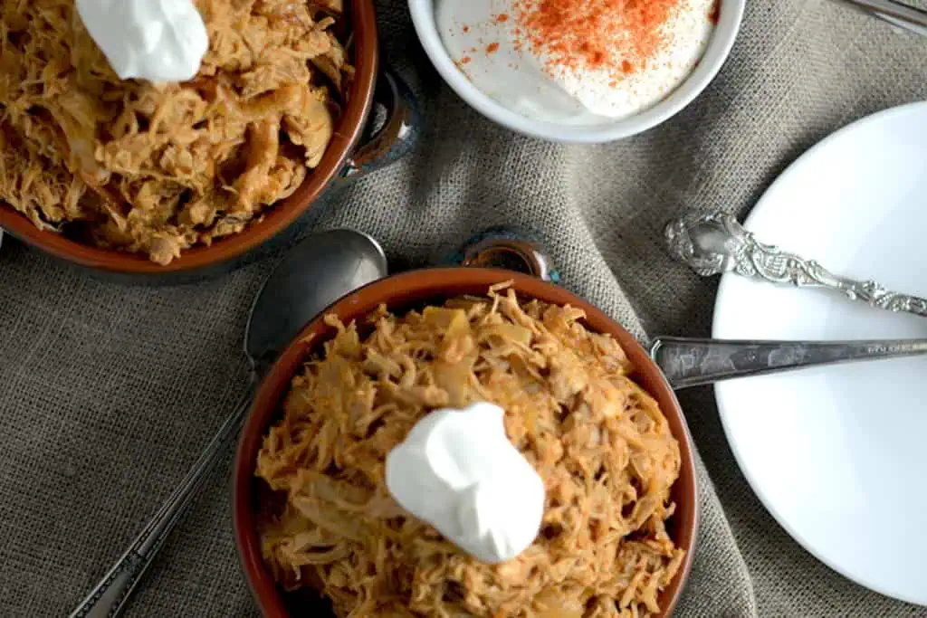 Overhead view of two bowls filled with shredded chicken paprikash, each topped with sour cream. A small bowl of paprika-seasoned sour cream is next to the bowls, along with silverware.