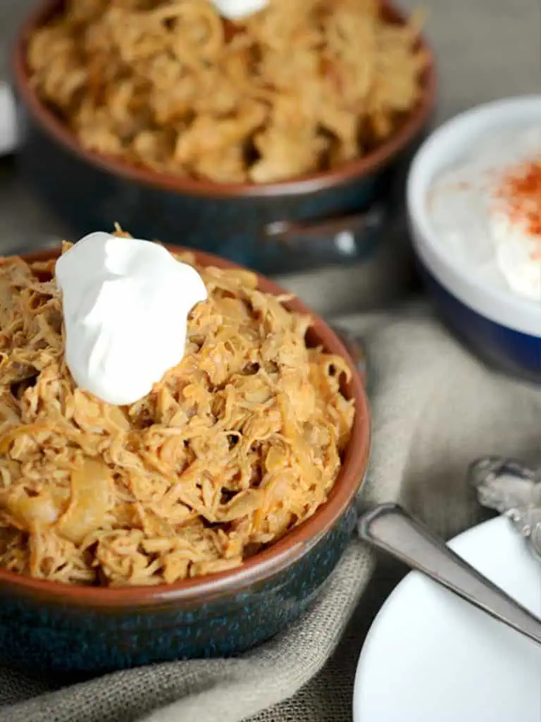 A close-up of a bowl filled with shredded chicken paprikash, topped with a dollop of sour cream. Another bowl of the dish is visible in the background.