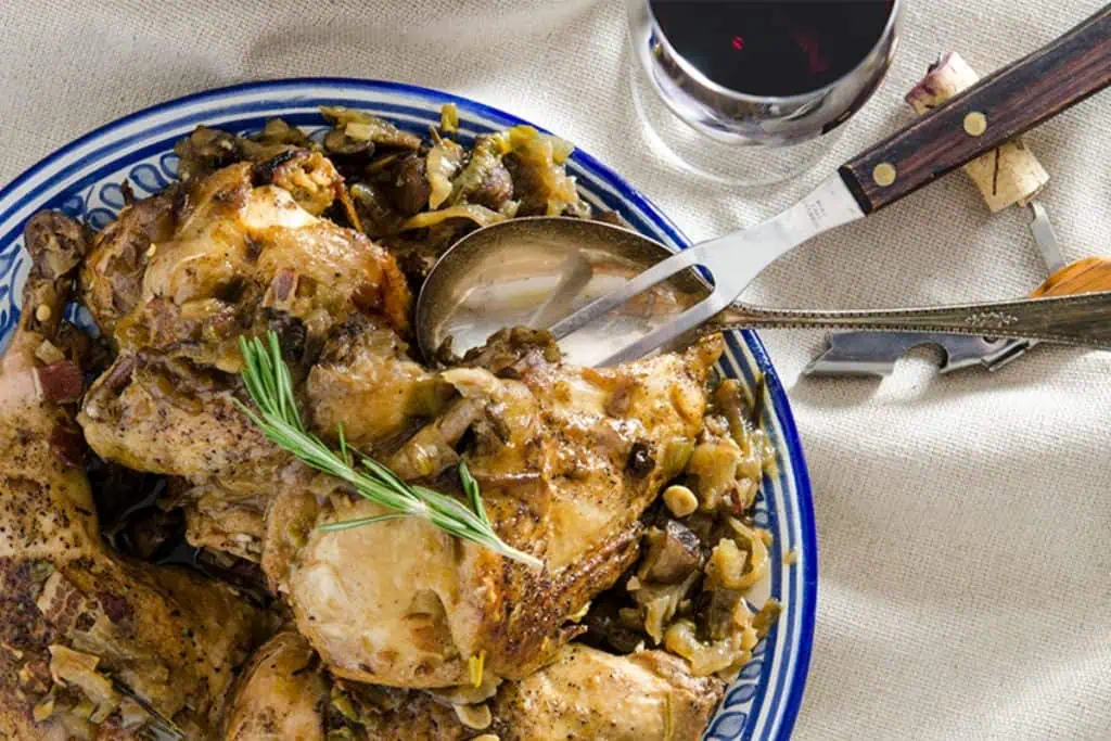 A plate of Slow-Cooker Coq au Vin garnished with a sprig of rosemary, with a glass of red wine and stacked plates in the background.