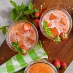 Top view of three glasses filled with strawberry lemonade, garnished with fresh strawberries and mint leaves, on a wooden board with a green checkered cloth.