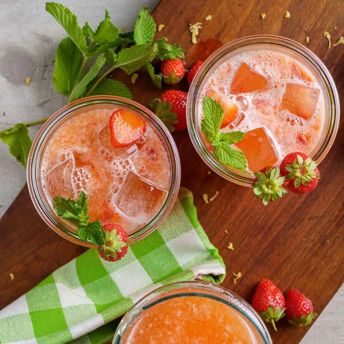 Top view of three glasses filled with strawberry lemonade, garnished with fresh strawberries and mint leaves, on a wooden board with a green checkered cloth.