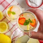 Hand holding a glass of sugar-free lemonade garnished with lemon slices, strawberries, and fresh mint leaves. A red and white checkered cloth, fresh lemons, and a pitcher of lemonade are in the background.