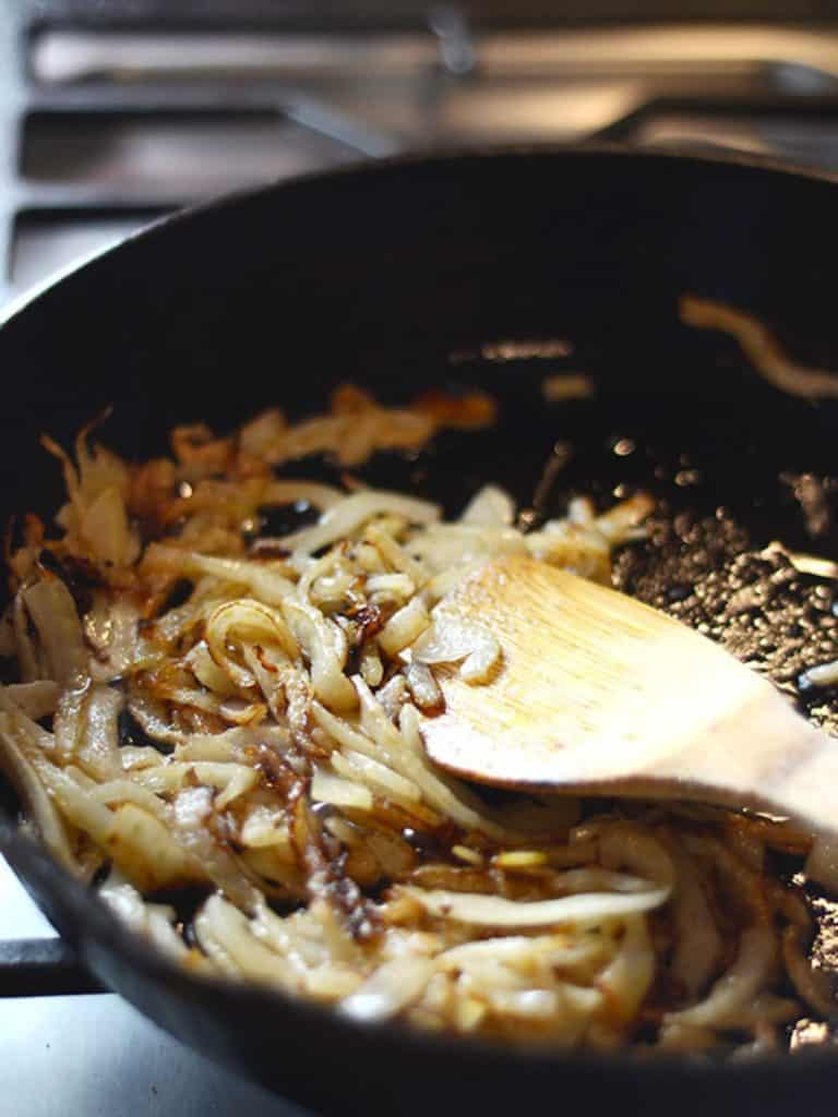 Onions being caramelized in a black skillet with a wooden spoon.
