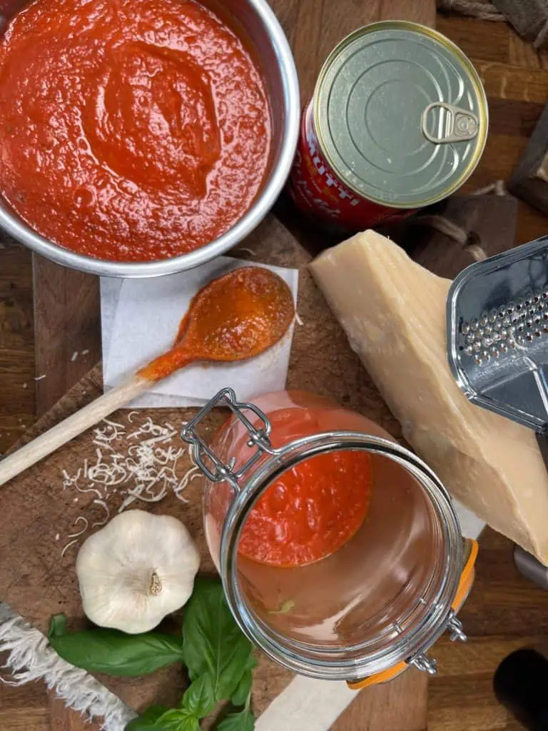 An overhead view of a jar being filled with homemade tomato sauce, placed on a wooden board with a spoon of sauce, a can of tomatoes, a wedge of Parmesan cheese, a garlic bulb, and fresh basil leaves.