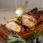 Overhead view of a whole bacon-wrapped pork tenderloin resting on a wooden cutting board with fresh herbs and a knife beside it.