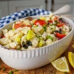 A close-up of a white ceramic dish filled with a vibrant cauliflower salad, featuring chopped cherry tomatoes, black olives, yellow bell peppers, and chives. The salad is placed on a wooden surface, with lemon wedges nearby and a blue-checkered cloth in the background.