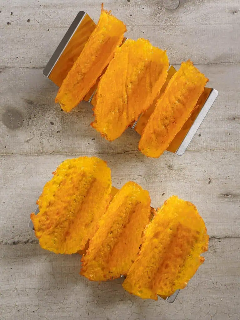 Overhead view of cheese taco shells in a metal holder on a light wooden surface, showcasing their texture and color.