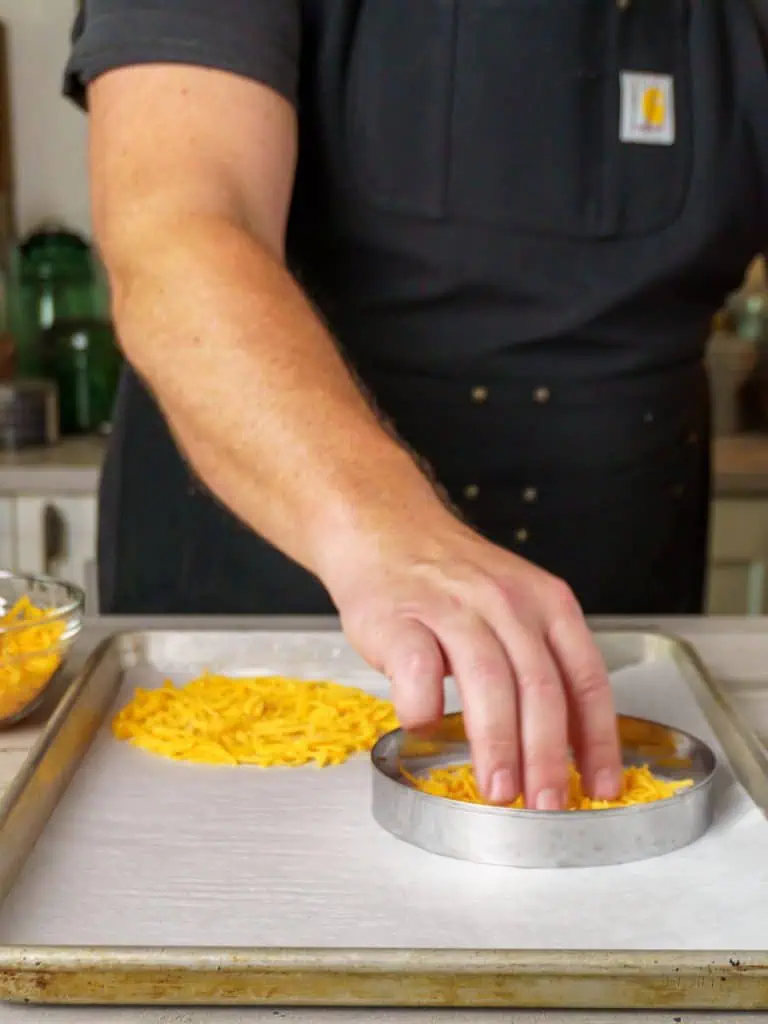 A person wearing a black apron placing shredded cheddar cheese inside the metal ring on the baking sheet.