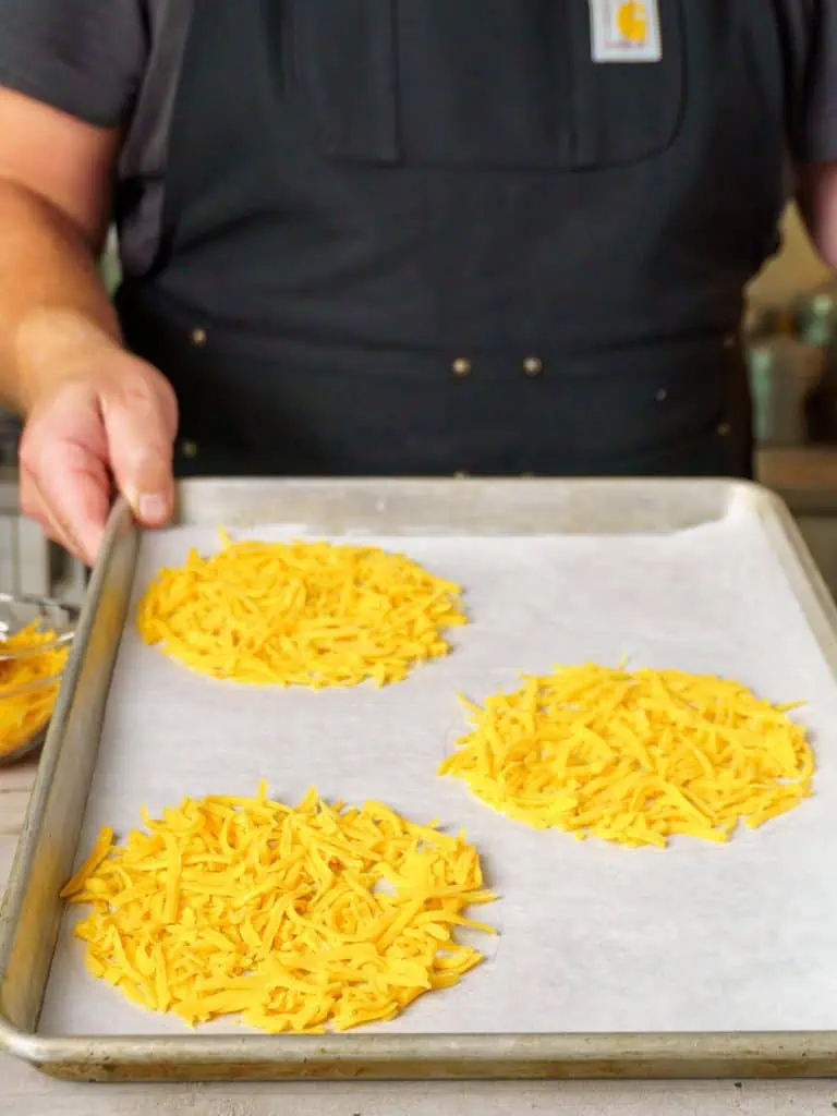 Three mounds of shredded cheddar cheese arranged on a parchment-lined baking sheet, ready to be baked.