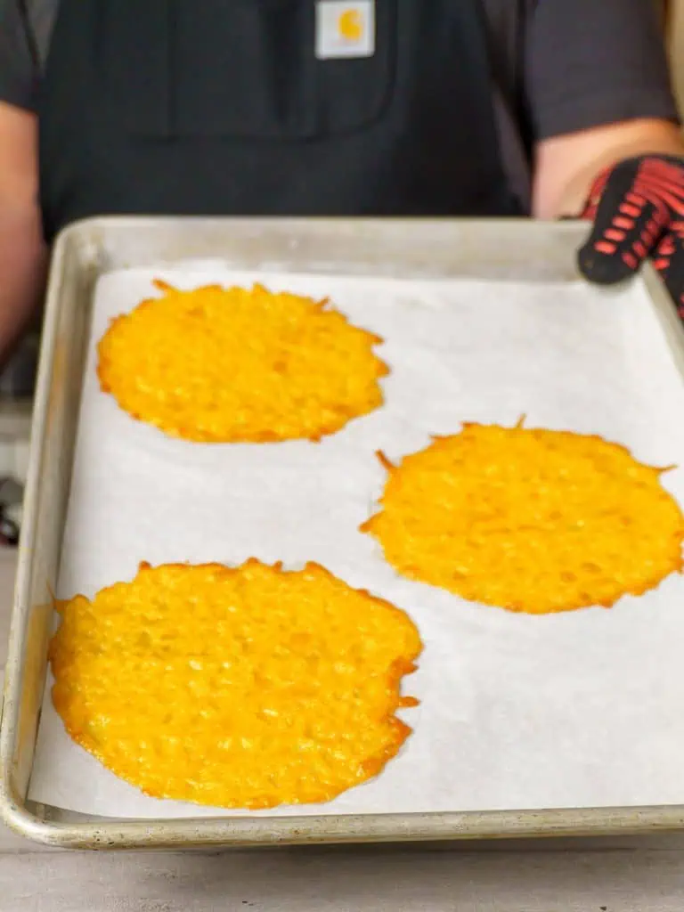 A person wearing a black apron holding a baking sheet with three melted and slightly crisp cheese circles.
