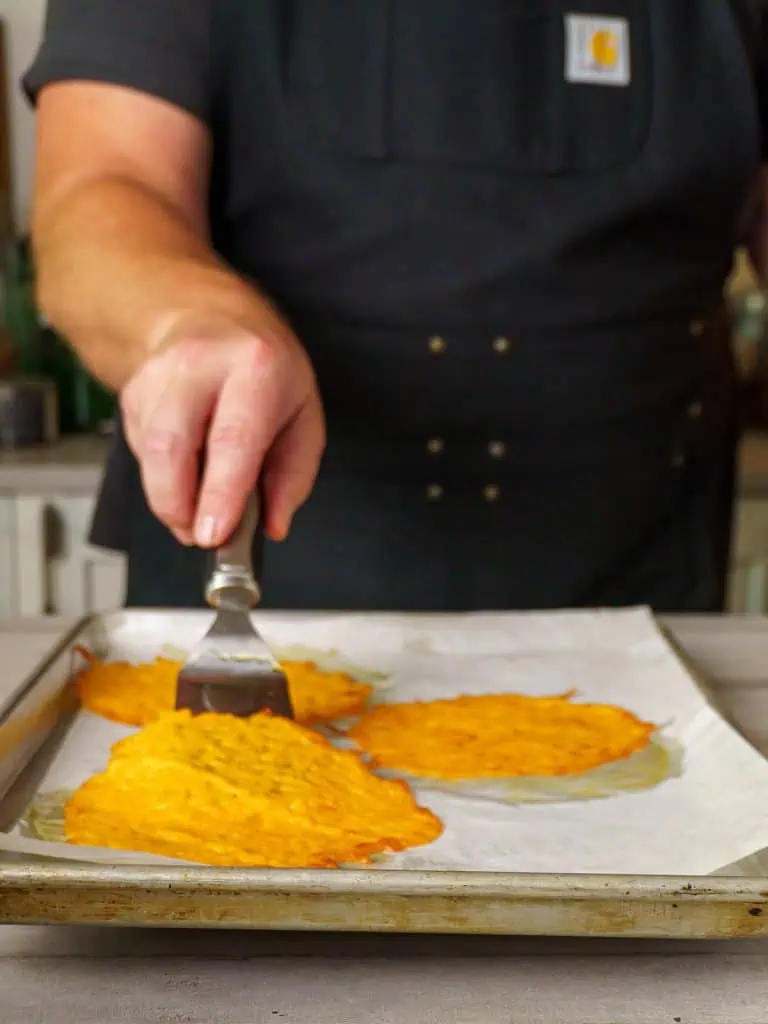 A person using a spatula to lift a melted cheese circle from the parchment-lined baking sheet.