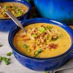 A close-up of two bowls of cheeseburger soup in blue ceramic bowls, topped with bacon and green onions, with a large blue pot in the background.
