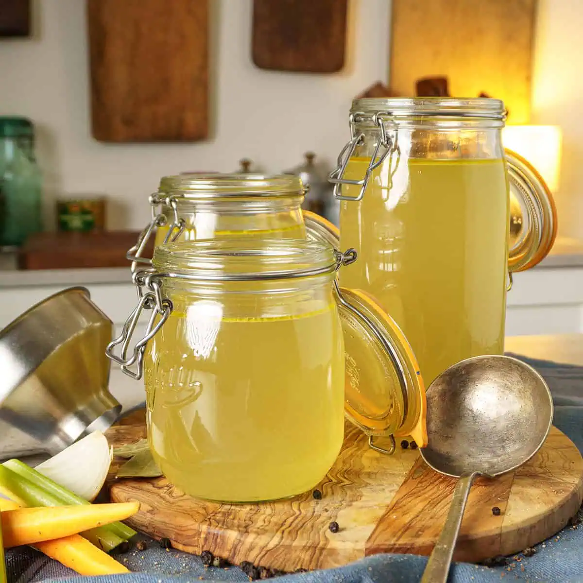 Three jars of homemade chicken broth with a metal ladle and kitchen funnel, all on a wooden cutting board in a kitchen setting.