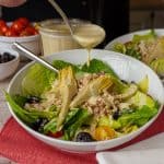A close-up shot of a fresh chicken Caesar salad in a white bowl. The salad is topped with shredded chicken, artichoke hearts, black olives, and cherry tomatoes. A hand is shown pouring creamy dressing over the salad. In the background, there is a jar of the dressing and a bowl of cherry tomatoes.