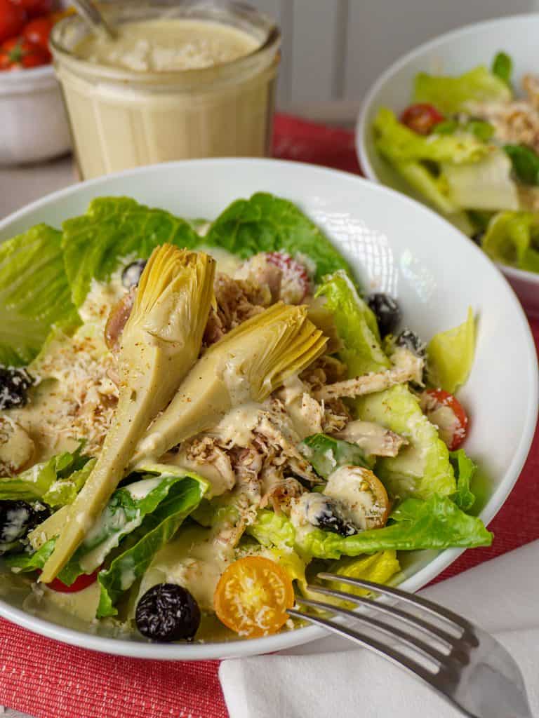 A close-up view of a chicken Caesar salad in a white bowl, featuring shredded chicken, artichoke hearts, black olives, and cherry tomatoes. A jar of creamy dressing is visible in the background. The salad is placed on a red cloth, and a white napkin with a fork is partially visible.
