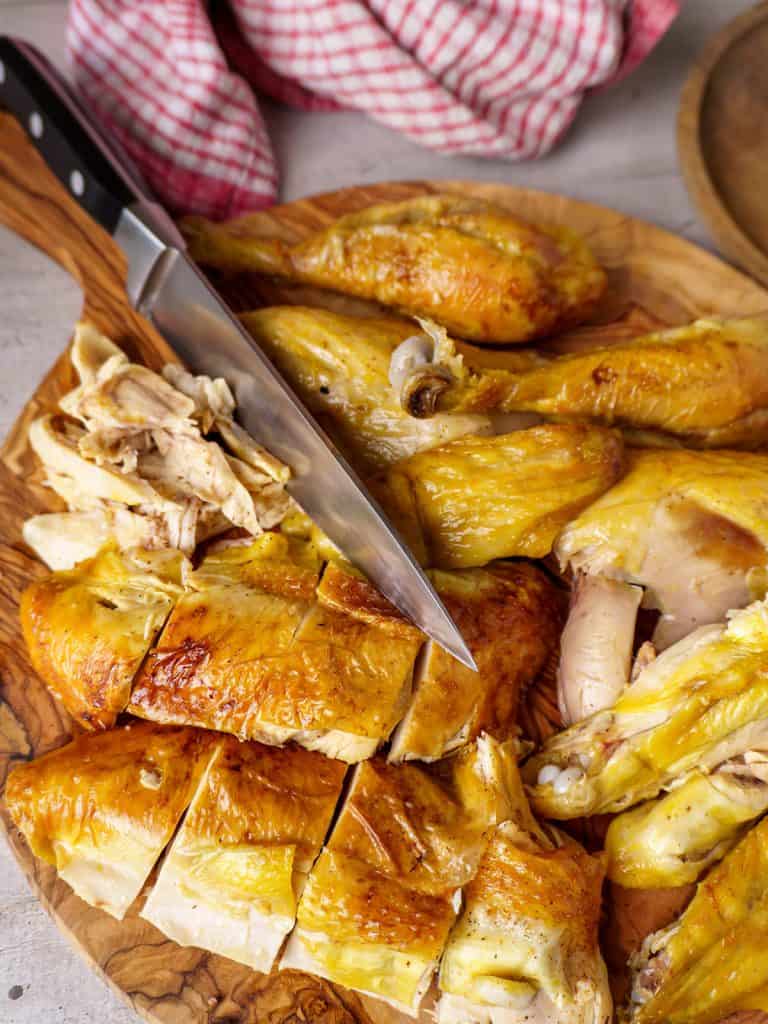 Close-up of a sliced rotisserie chicken on a wooden cutting board, showing golden-brown skin and juicy meat, with a knife placed beside the chicken.
