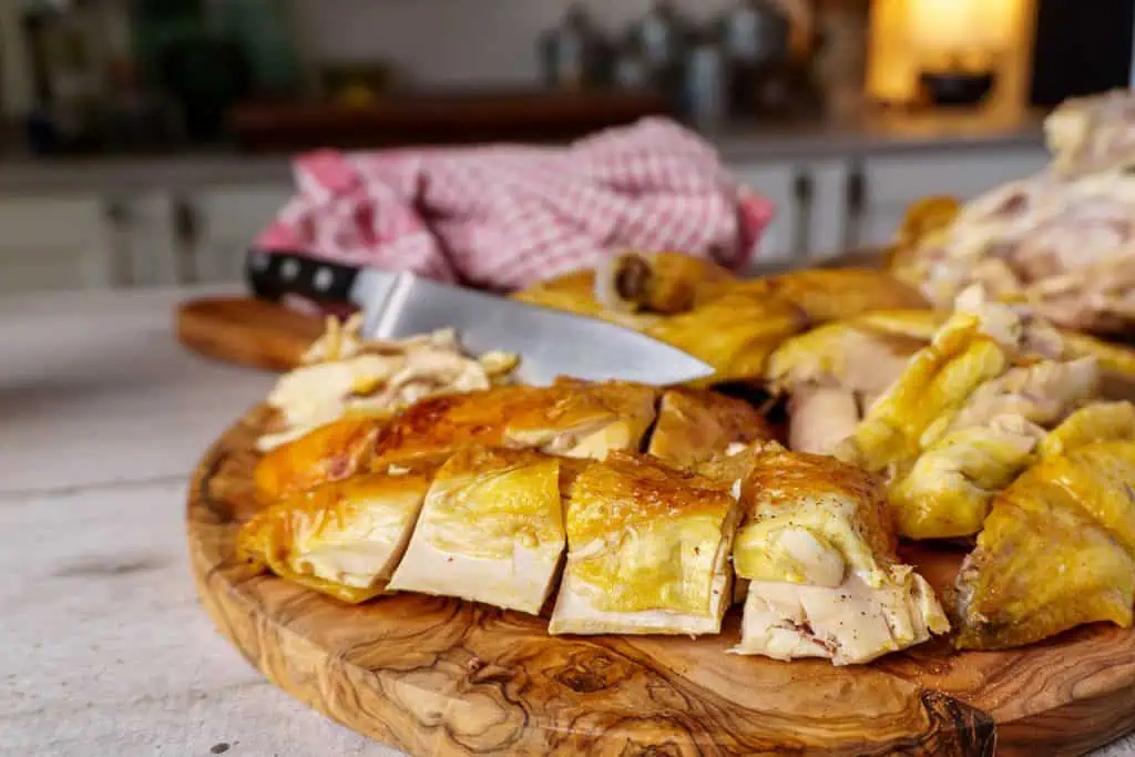 A horizontal view of a sliced rotisserie chicken on a wooden cutting board, showcasing the juicy, golden skin, with a knife and checkered cloth in the background.