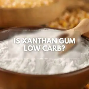 A close-up of a bowl of xanthan gum with a wooden spoon in the background, featuring the text "Is Xanthan Gum Low Carb?" overlaying the center of the image.