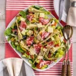Overhead view of a fresh Italian salad topped with slices of cured meat, artichoke hearts, olives, and shaved Parmesan cheese, set on a red and white striped cloth with cutlery on the side.