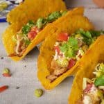 Close-up of rotisserie chicken tacos with cheddar cheese shells filled with lettuce, tomatoes, and cilantro, resting on a wooden table with toppings scattered around.