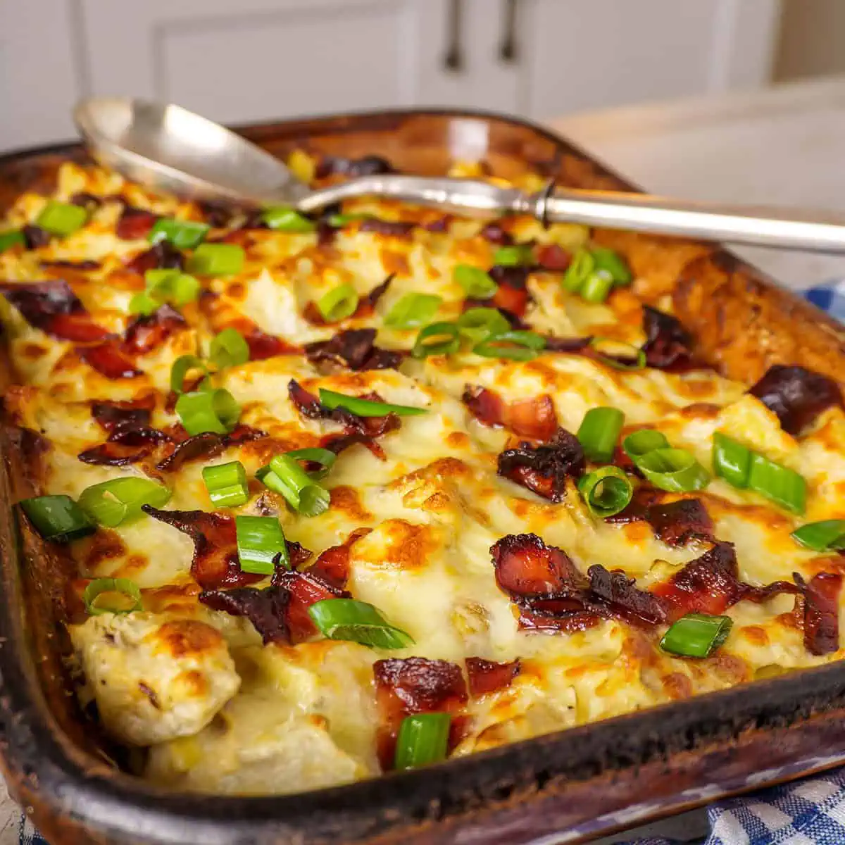 A close-up of a cheesy baked rotisserie chicken casserole topped with crispy bacon and fresh green onions, with a serving being lifted by a fork.