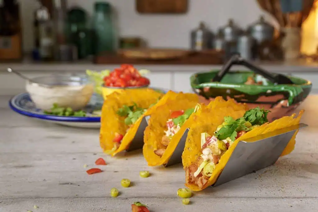 Rotisserie chicken tacos served in cheese shells, lined up on a taco holder with bowls of toppings like diced tomatoes and shredded lettuce in the background on a rustic kitchen counter.
