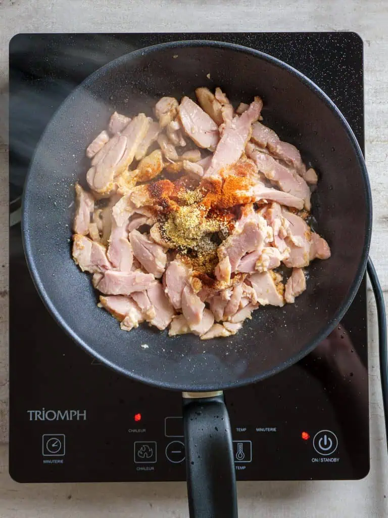 Raw shredded rotisserie chicken in a pan with various spices added on top, ready to be cooked on an induction cooktop.