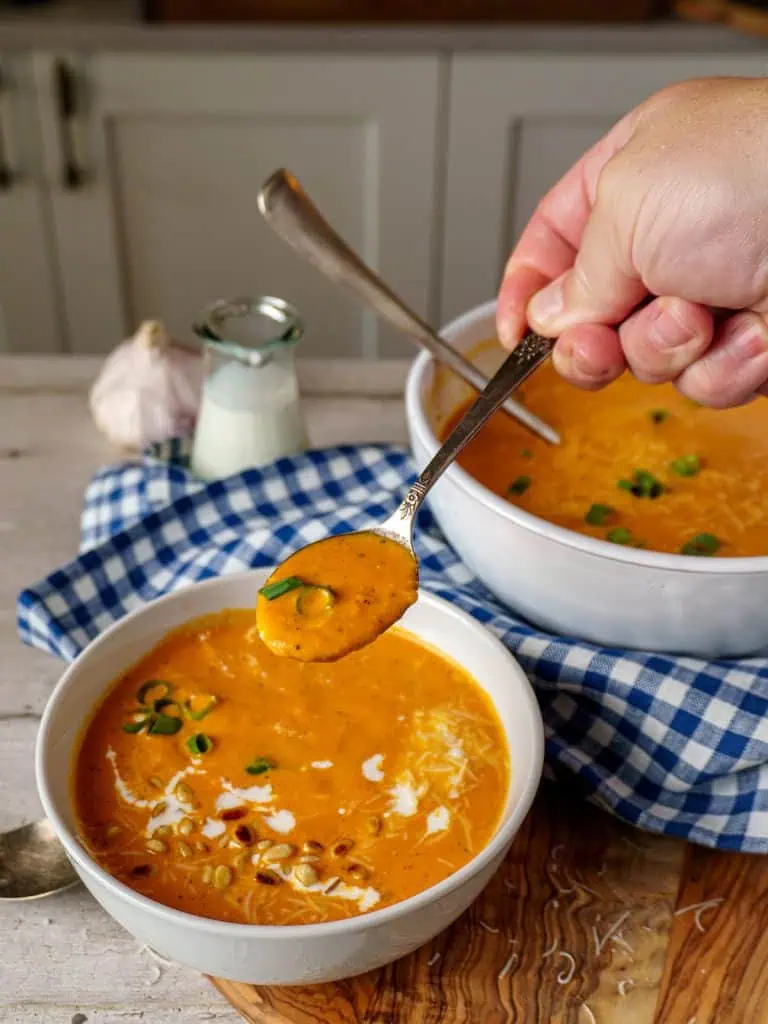 A close-up of a hand holding a spoonful of creamy tomato bisque soup, hovering over a white bowl filled with the soup, garnished with green onions and pine nuts. A larger bowl of soup and a checkered blue cloth are visible in the background.