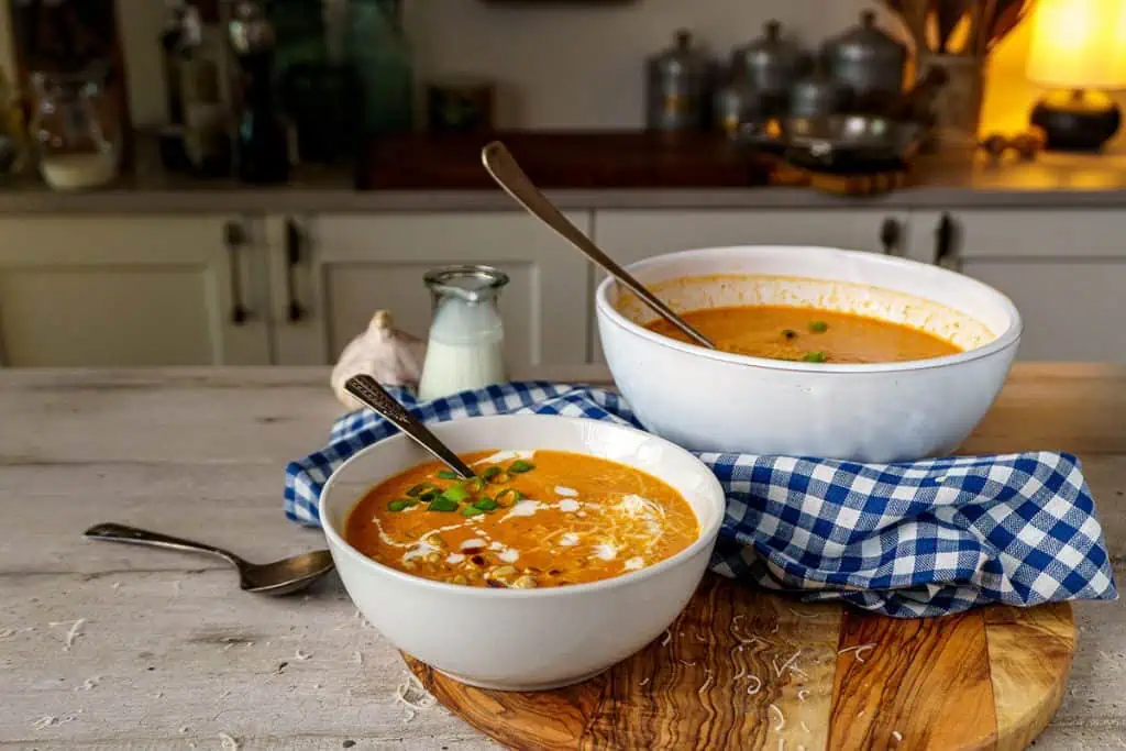Two white bowls of creamy tomato bisque soup, one in the foreground and one in the background, both garnished with green onions and pine nuts. The setting includes a rustic kitchen background, a blue checkered cloth, and a garlic bulb.