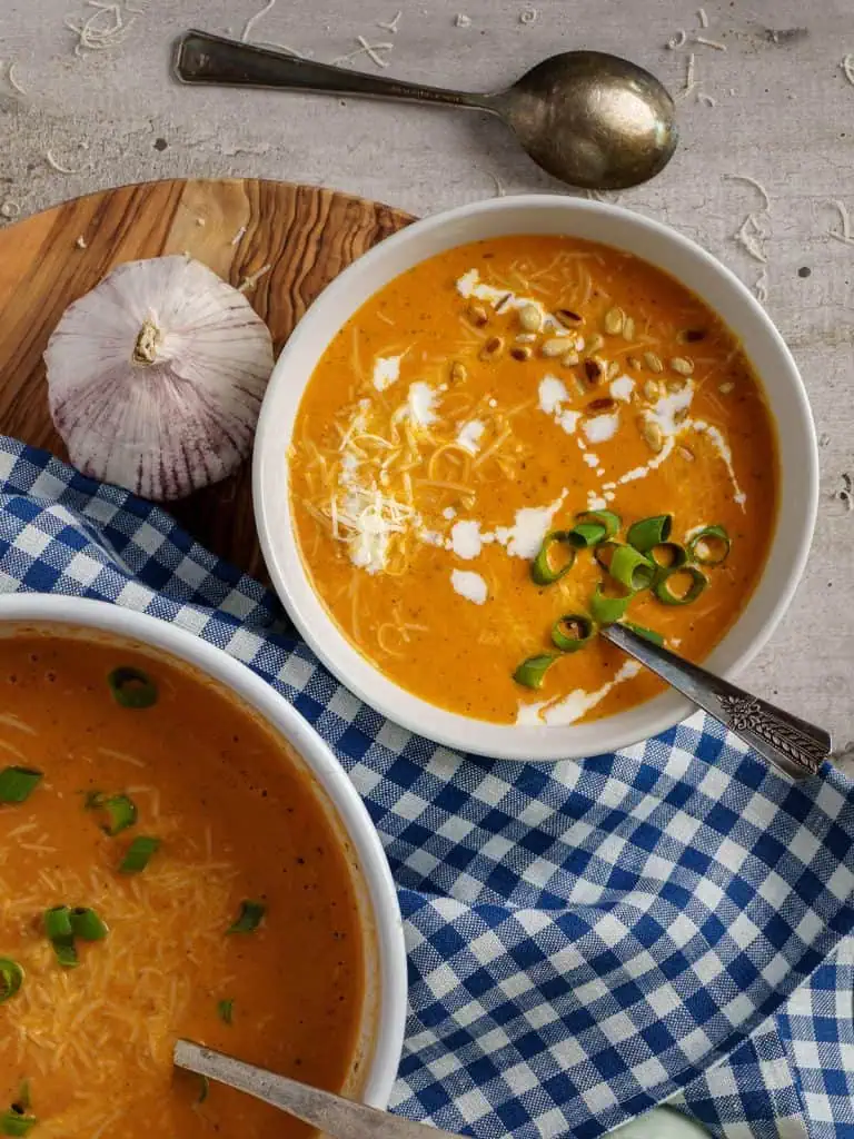 An overhead view of two white bowls of creamy tomato bisque soup, garnished with green onions, pine nuts, and a swirl of cream. A whole garlic bulb and a blue checkered cloth are arranged on a wooden surface beside the bowls.