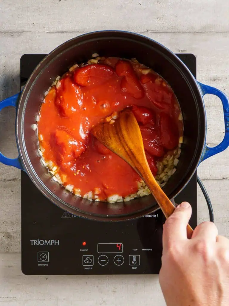 Adding canned tomatoes to the sautéed onions in the pot.