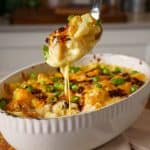 A spoonful of cheesy cauliflower mac and cheese being lifted from a baked dish, topped with melted cheese, crispy bacon, and garnished with chopped green onions.