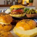 A juicy hamburger served on a golden, fluffy keto hamburger bun, topped with fresh slices of tomato, red onion, and greens, with a side of pickles. Additional buns with sesame seeds are placed around the scene on a rustic wooden board.