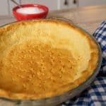 Golden-brown keto pie crust baked and ready in a glass pie dish with a red bowl of sugar in the background, next to a blue and white checkered cloth.
