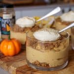 A glass cup of pumpkin cheesecake topped with whipped cream and spiced crumbles, with a small pumpkin and jar of pumpkin spice in the background, placed on a wooden board.
