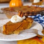 Close-up side view of a pumpkin pie slice with a golden crust, topped with whipped cream and a pecan, served on a plate with a fork. The whole pumpkin pie is visible in the background.