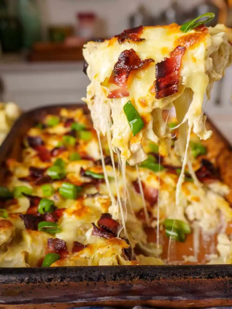 Close-up of a serving spoon dipping into a creamy, golden-brown rotisserie chicken casserole, with melted cheese, crispy bacon, and garnished with green onions.