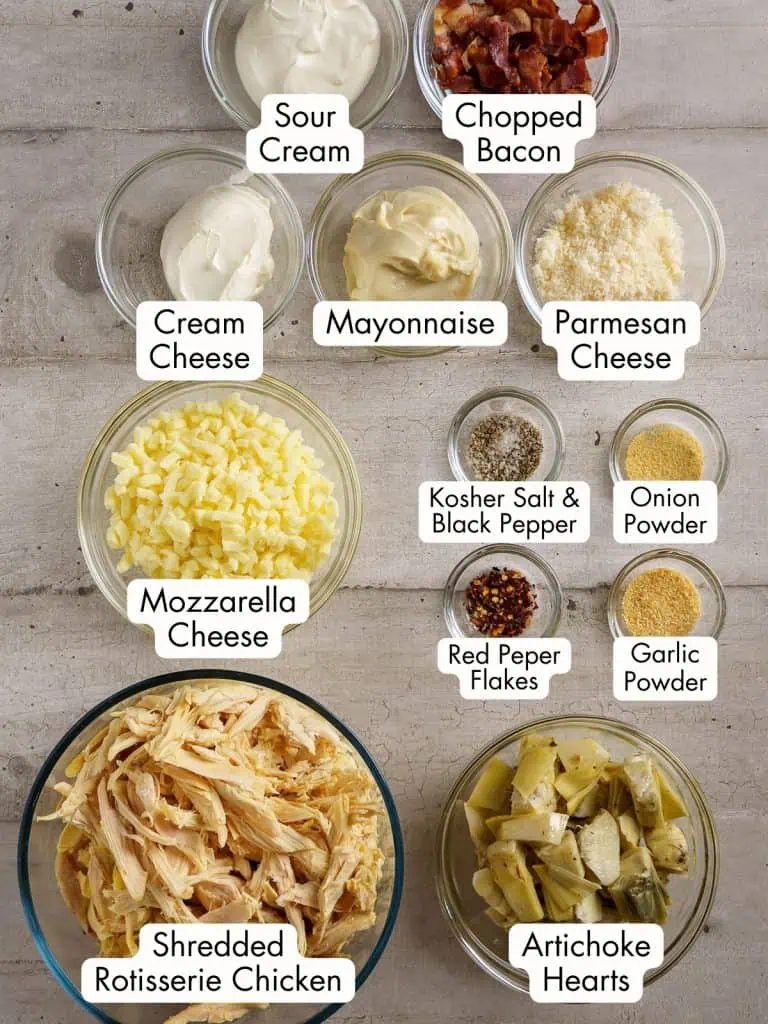 Bowl of ingredients for a chicken casserole, labeled from top to bottom: sour cream, chopped bacon, cream cheese, mayonnaise, Parmesan cheese, mozzarella cheese, kosher salt and black pepper, red pepper flakes, onion powder, garlic powder, shredded rotisserie chicken, and artichoke hearts.