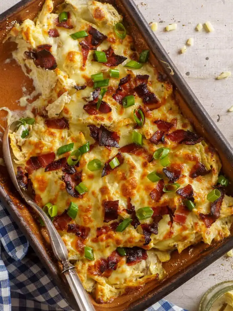 Overhead view of a baked rotisserie chicken casserole with layers of golden-brown cheese, crispy bacon, and green onions, placed on a blue-checkered napkin.