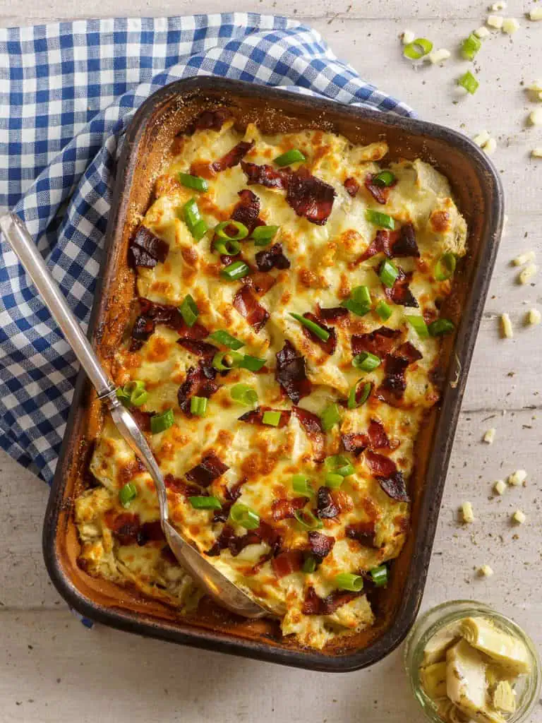 Overhead shot of a rotisserie chicken casserole in a baking dish, featuring bubbly cheese, crispy bacon, and green onions, with a spoon resting on the side, ready to serve.