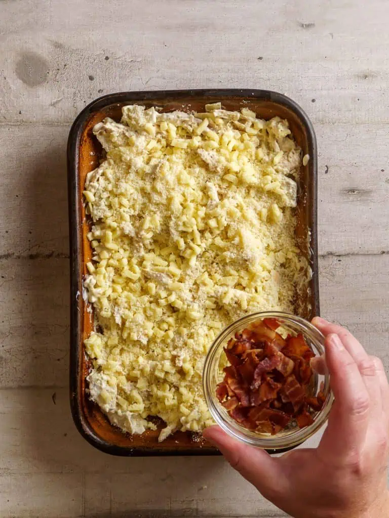 Chopped bacon being sprinkled on top of the shredded cheese in the casserole dish.