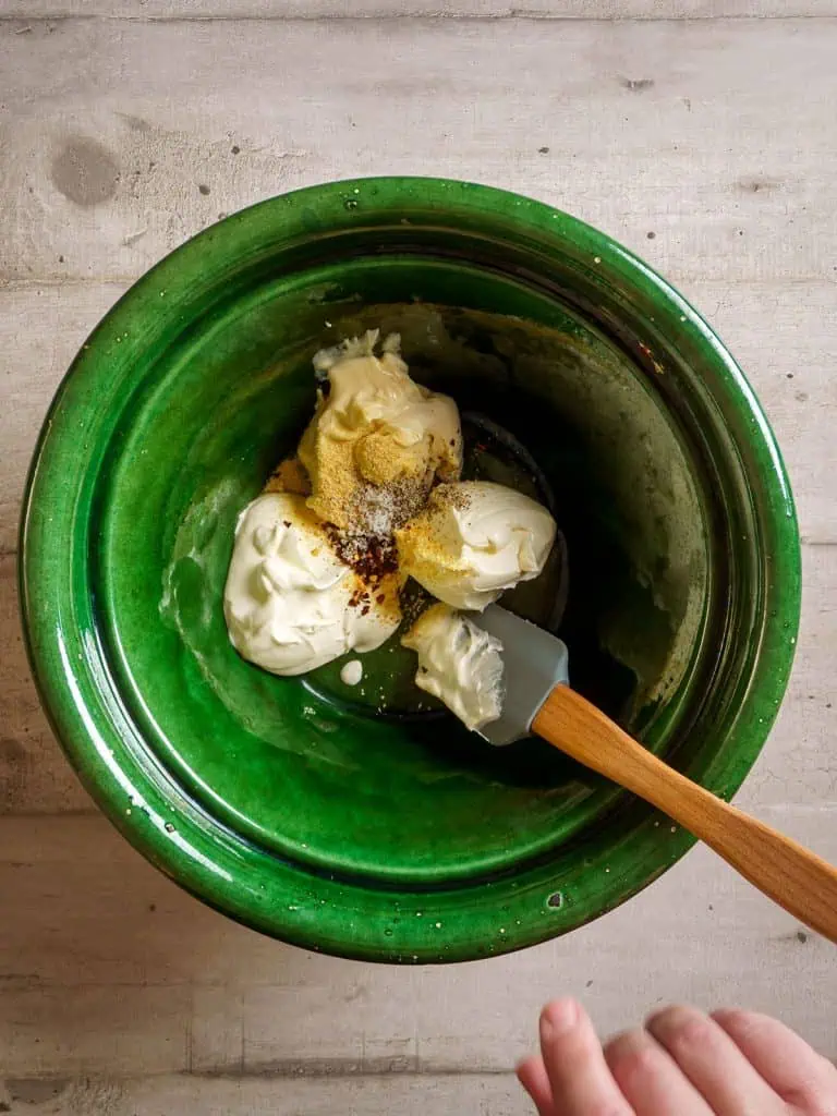 Sour cream, cream cheese, mayonnaise, and spices in a green bowl with a spatula, ready to be mixed.