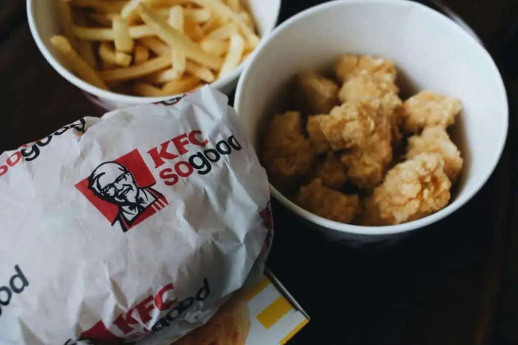 Close-up of a KFC meal including fries and chicken bites in bowls, with a wrapped sandwich in the foreground, displaying KFC's "So Good" branding.