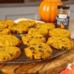 Close-up of a rack filled with pumpkin chocolate chip cookies, golden-brown and dotted with dark chocolate chips. A jar of pumpkin pie spice and a decorative pumpkin are visible in the background.